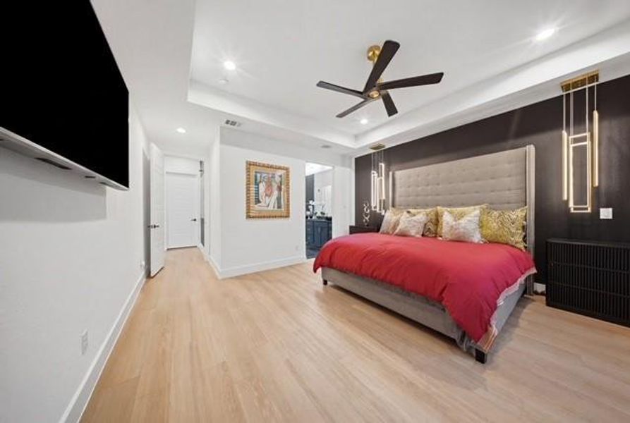 Spacious Primary bedroom featuring light wood-finish flooring, a tray ceiling with recessed lighting, a ceiling fan, and modern wall-mounted light fixtures flanking the bed