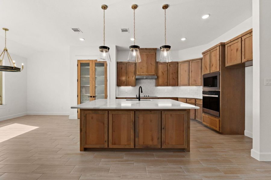 Kitchen with brown cabinets, pendant lighting, backsplash, stainless steel appliances, and an island with sink