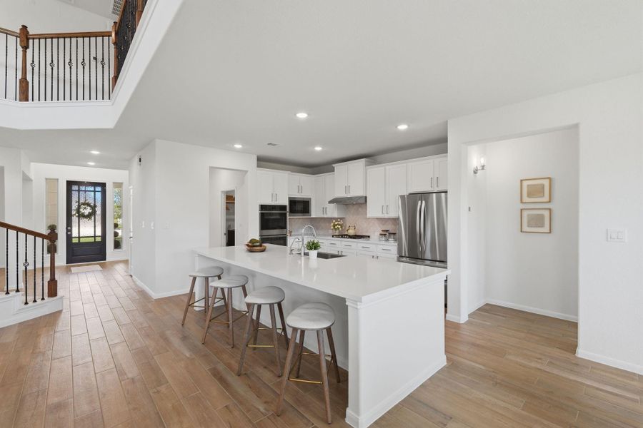 This photo showcases a spacious, modern kitchen with a large island, white cabinetry, stainless steel appliances, and bar seating. The open-concept area features hardwood floors and a view of the entrance, creating a welcoming and functional space.