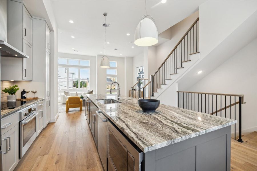 This modern kitchen features sleek cabinetry, a large island with a striking marble countertop, and pendant lighting. It opens into a bright living area with large windows and a cozy seating nook, complemented by a stylish staircase. Perfect for entertaining and everyday living.