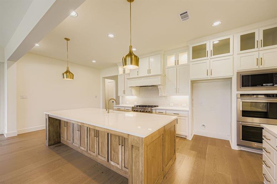 Kitchen featuring glass insert cabinets, white cabinets, decorative light fixtures, appliances with stainless steel finishes, and a center island with sink