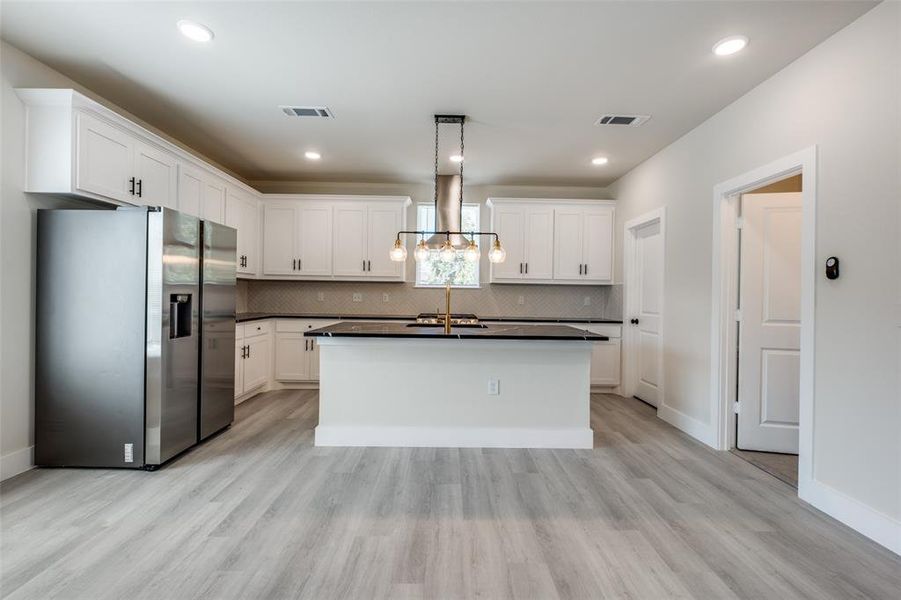 Kitchen featuring stainless steel fridge, a kitchen island with sink, decorative light fixtures, white cabinetry, and wall chimney exhaust hood