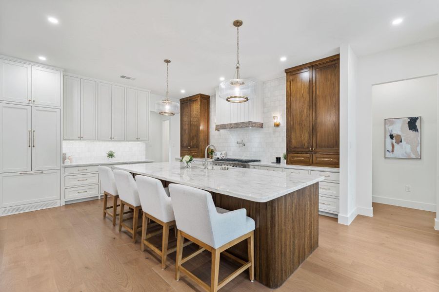 Designer kitchen with a massive island wrapped in rich wood tones and topped with stunning Carrara marble counters. Custom cabinetry, statement lighting, and a full-height tile backsplash create a high-end feel throughout.