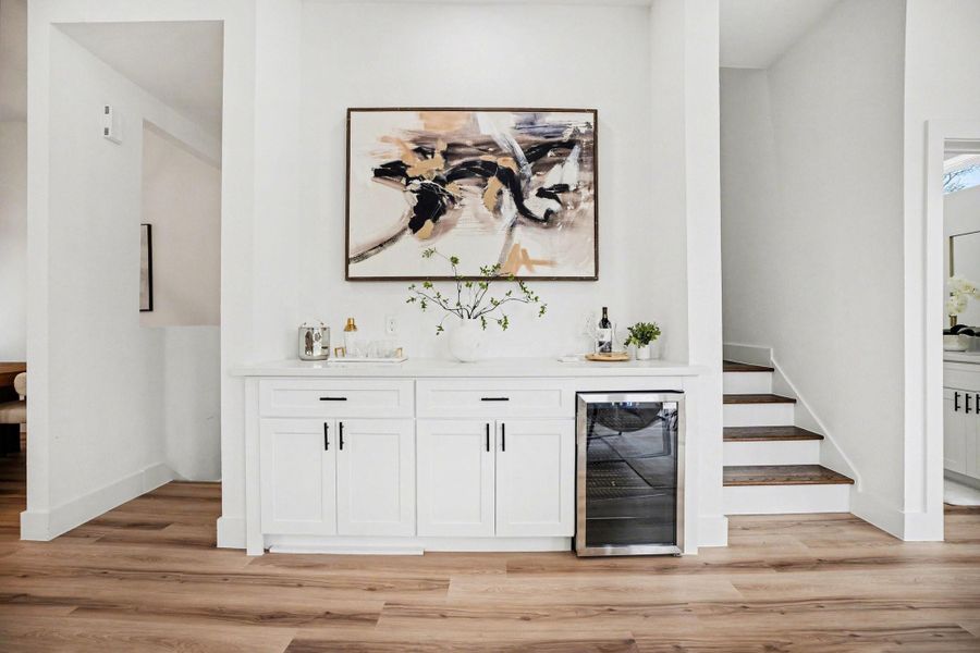 This photo showcases a stylish, modern interior with a built-in bar area. It features white cabinetry, a countertop with decorative items, and a wine cooler beneath.