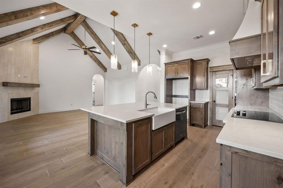 Open-concept living area featuring a kitchen island with a farmhouse sink, wood-finish flooring, vaulted ceilings with exposed beams, a fireplace with a wood mantel, and arched doorways