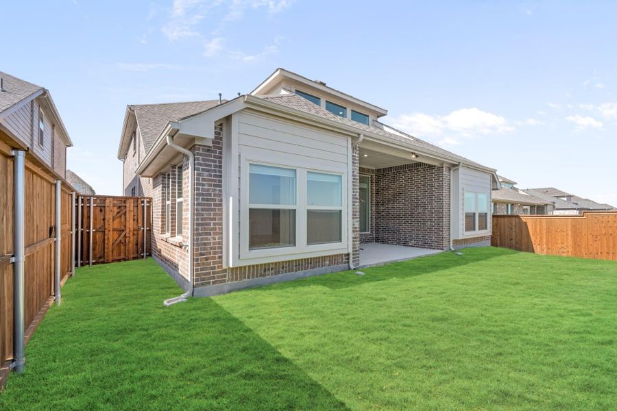 Rear exterior of brick home with covered patio, large windows, and fenced backyard with green lawn Rear exterior of brick home with covered patio, large windows, and fenced backyard with green lawn