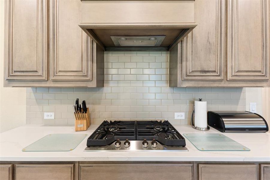 Kitchen featuring a stainless steel gas cooktop with four burners, a built-in range hood, light gray subway tile backsplash, light wood-finish cabinetry, and solid surface countertops Kitchen featuring a stainless steel gas cooktop with four burners, a built-in range hood, light gray subway tile backsplash, light wood-finish cabinetry, and solid surface countertops