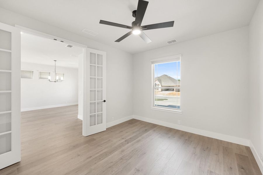 Empty room with hardwood floors, ceiling fan, and French doors opening to dining area with chandelier Empty room with hardwood floors, ceiling fan, and French doors opening to dining area with chandelier