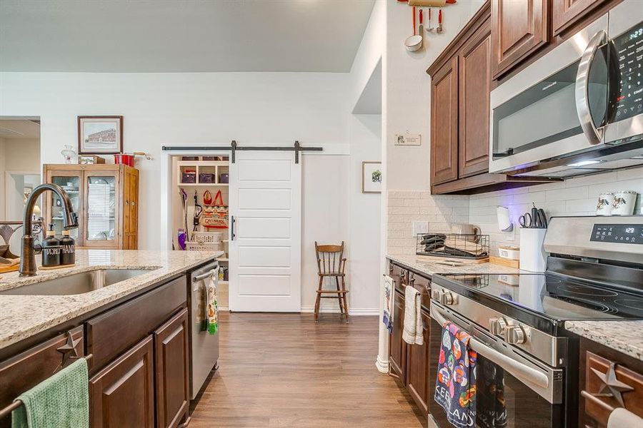 Kitchen featuring stainless steel appliances, a barn door, light stone countertops, dark brown cabinetry, and backsplash Kitchen featuring stainless steel appliances, a barn door, light stone countertops, dark brown cabinetry, and backsplash