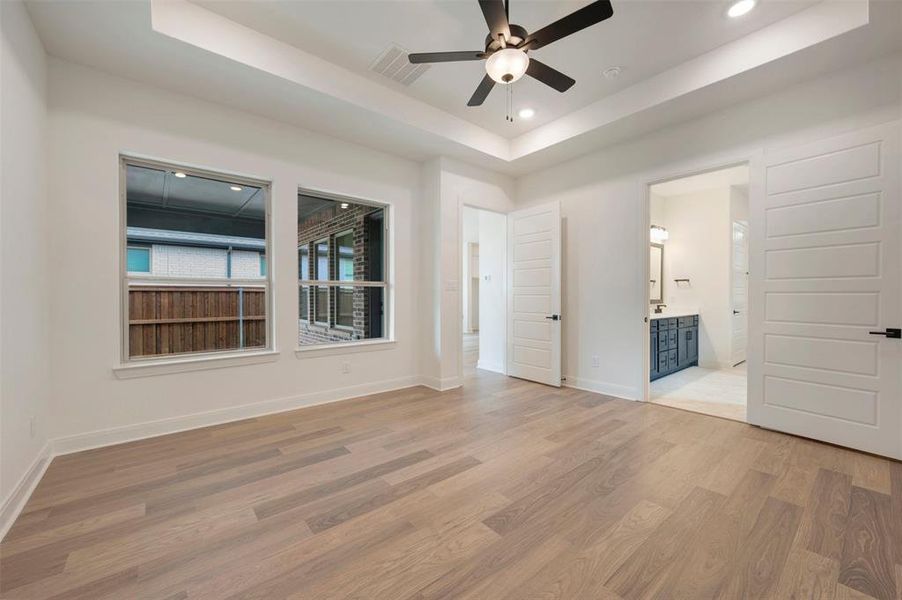 The spacious room features light wood flooring, white walls, and a tray ceiling with recessed lighting and a ceiling fan