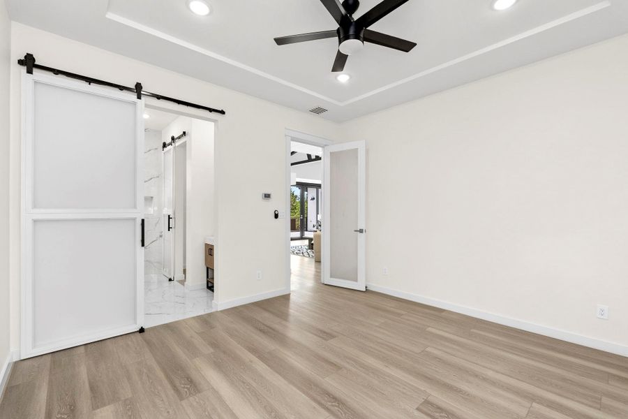 Unfurnished bedroom with a barn door, recessed lighting, ceiling fan, a tray ceiling, and light wood-style floors