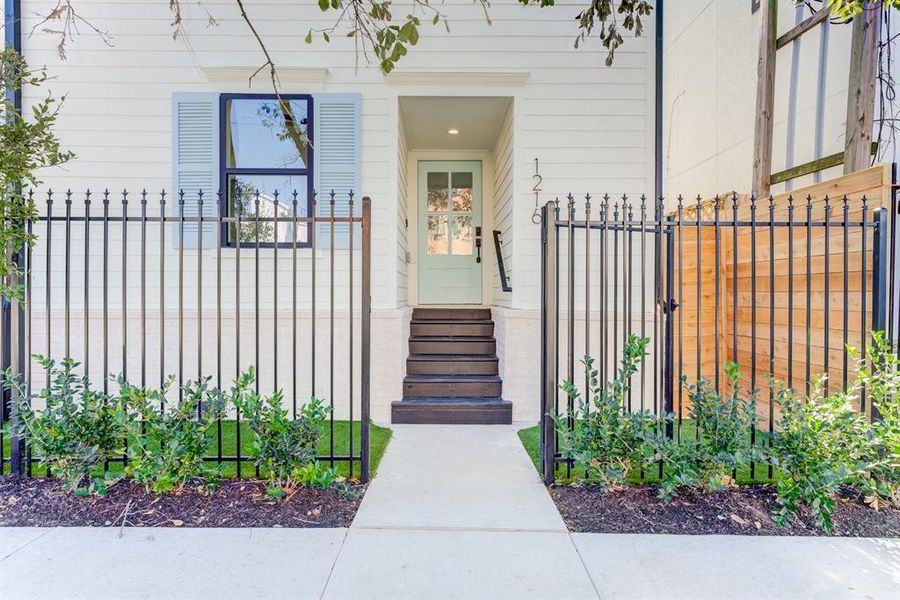 This photo showcases a modern fenced Victorian home entrance with a stylish door, black accented double paned windows, adjacent to a matching garage door. The exterior features clean white Hardie Plank siding neutral brick, and a small landscaped area with shrubs.