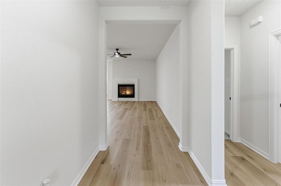 Hallway with wood-finish flooring extending into a living area featuring a fireplace with a white mantel and a ceiling fan