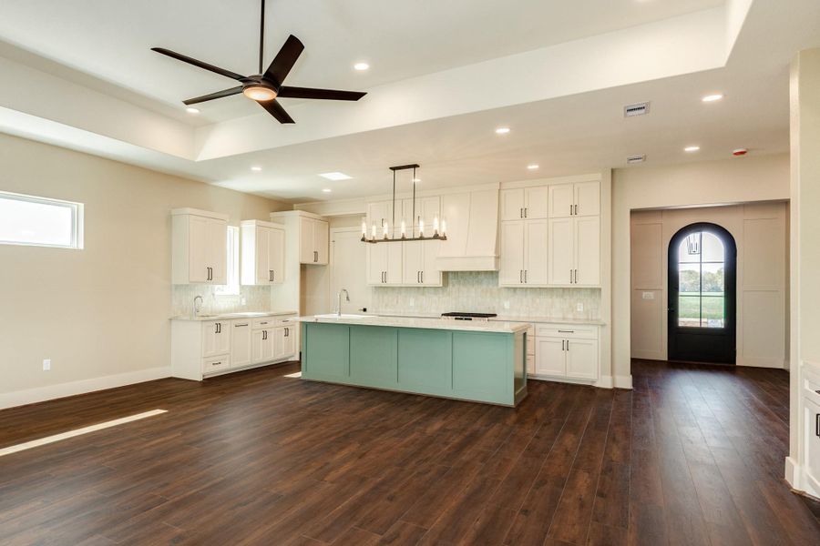 This spacious kitchen features modern custom cabinetry, a large island for entertaining, and elegant quartzite Taj Mahal countertops. The open layout is enhanced by dark wood flooring, recessed lighting, a ceiling fan, and a stylish light fixture. This space creates a warm and inviting space.