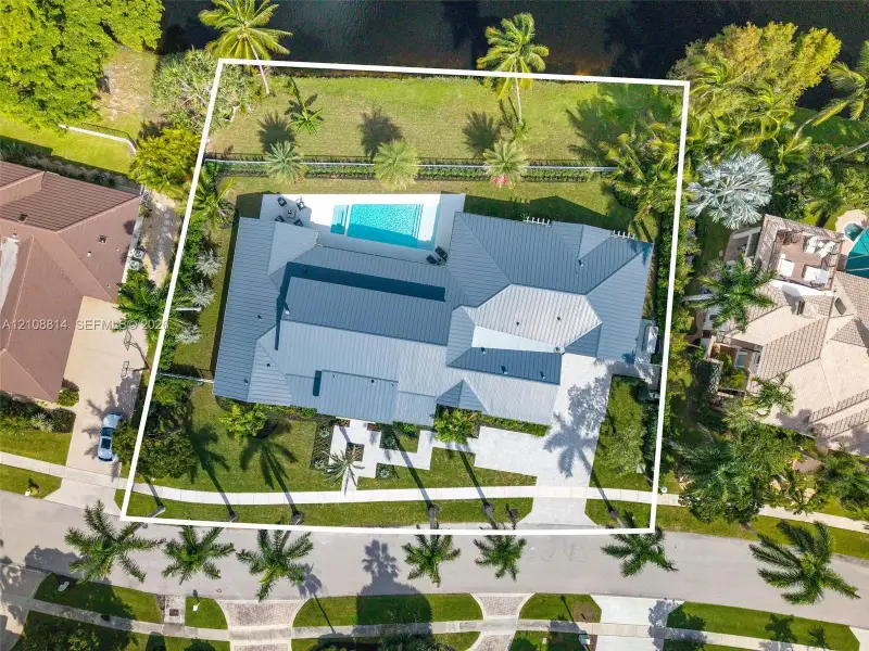 Aerial view of a residential property featuring a metal roof, an in-ground swimming pool, a paver driveway, and mature landscaping Aerial view of a residential property featuring a metal roof, an in-ground swimming pool, a paver driveway, and mature landscaping