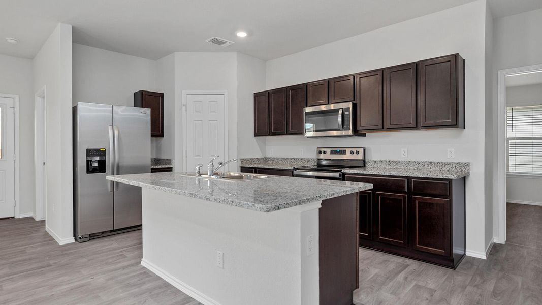 Kitchen with stainless steel appliances, dark wood finish cabinetry, a kitchen island with sink, light stone counters, and light wood-type flooring