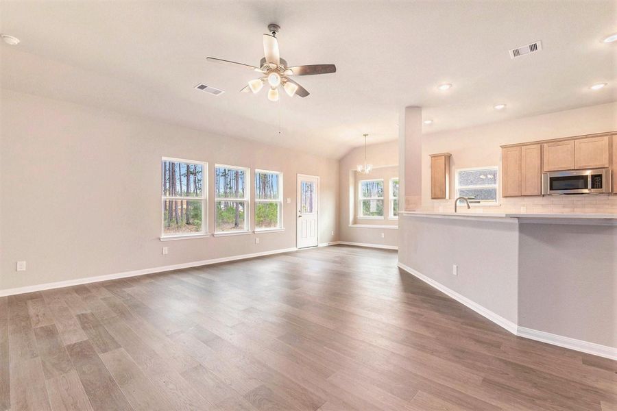 This spacious, open-concept living area features hardwood floors, abundant natural light from multiple windows, and a modern kitchen with light wood cabinets and stainless steel appliances. The room is enhanced by recessed lighting and a ceiling fan, creating a warm and inviting atmosphere.