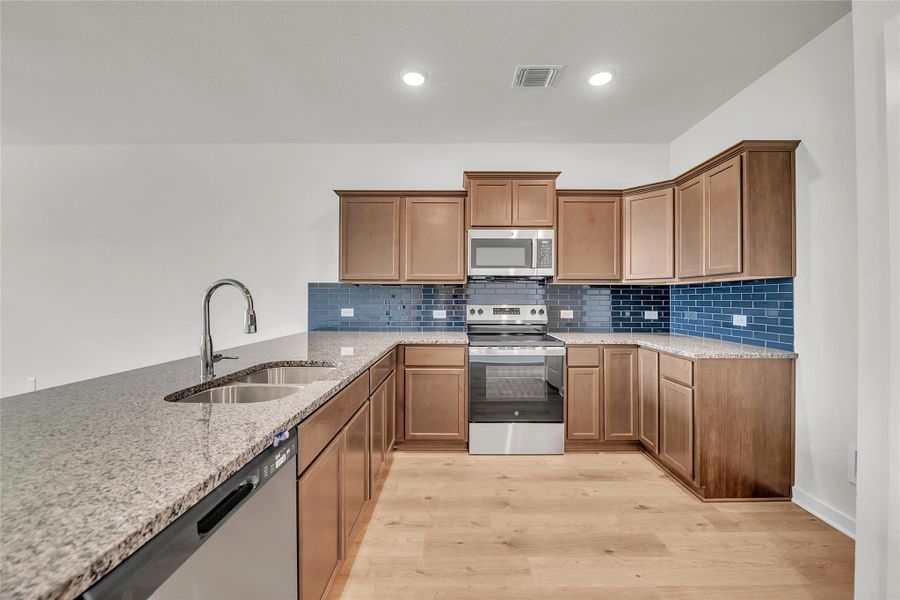 Kitchen featuring light stone counters, stainless steel appliances, decorative backsplash, light wood finished floors, and brown cabinets Kitchen featuring light stone counters, stainless steel appliances, decorative backsplash, light wood finished floors, and brown cabinets