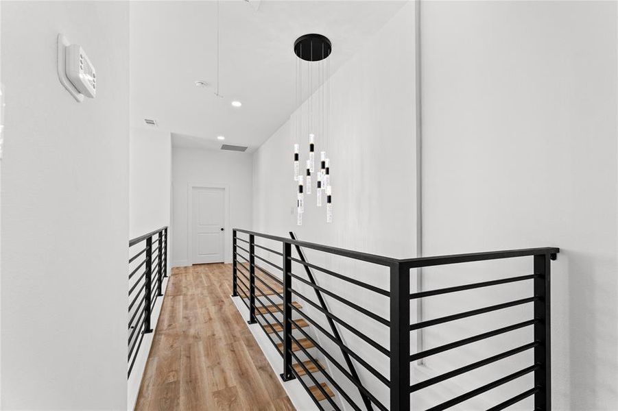 Upper-level hallway featuring wood-finish flooring, white walls, black metal railings, and a modern chandelier