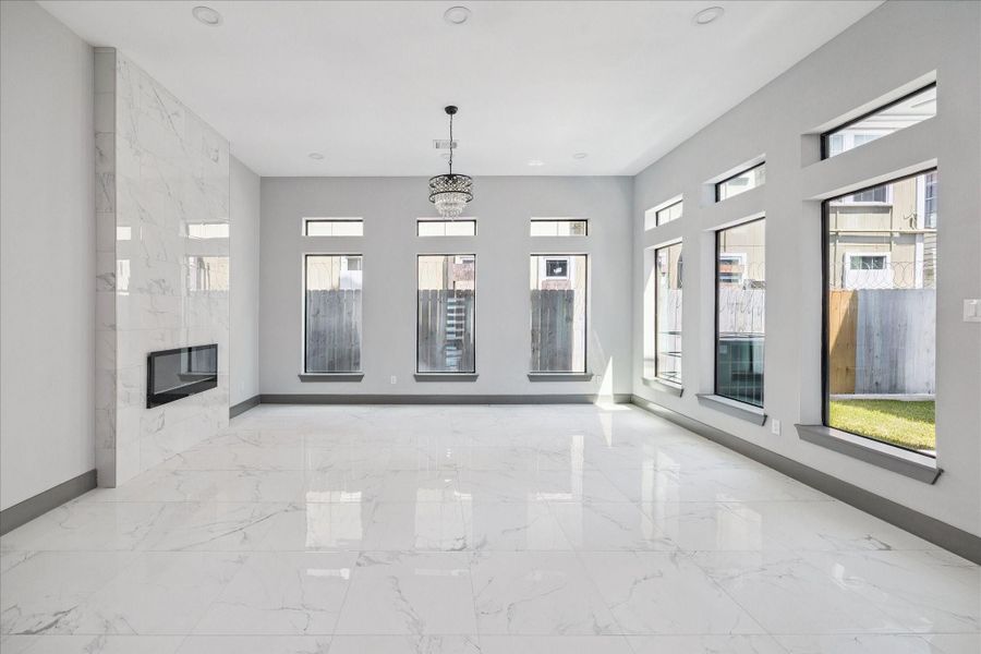 Expansive windows wrap two sides of the room, bringing in natural light and framing views of the yard. A full-height marble-look tile fireplace anchors the space with modern elegance, complemented by a crystal-accented chandelier that adds just the right touch of sophistication.
