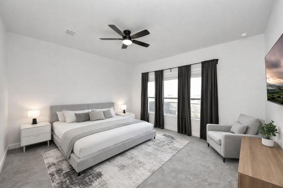 This spacious bedroom offers a serene retreat with elegant bedding, plush carpeting, and abundant natural light from expansive windows. A ceiling fan and cozy seating area complete the space, creating a relaxing and inviting environment.