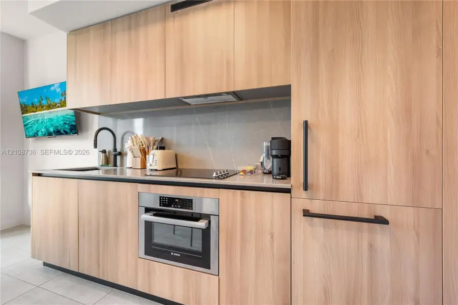 Modern cabinetry with a light wood-finish, sleek stone countertops, integrated stainless-steel wall oven, matte black hardware, and an undermount sink with a matching matte black faucet Modern cabinetry with a light wood-finish, sleek stone countertops, integrated stainless-steel wall oven, matte black hardware, and an undermount sink with a matching matte black faucet
