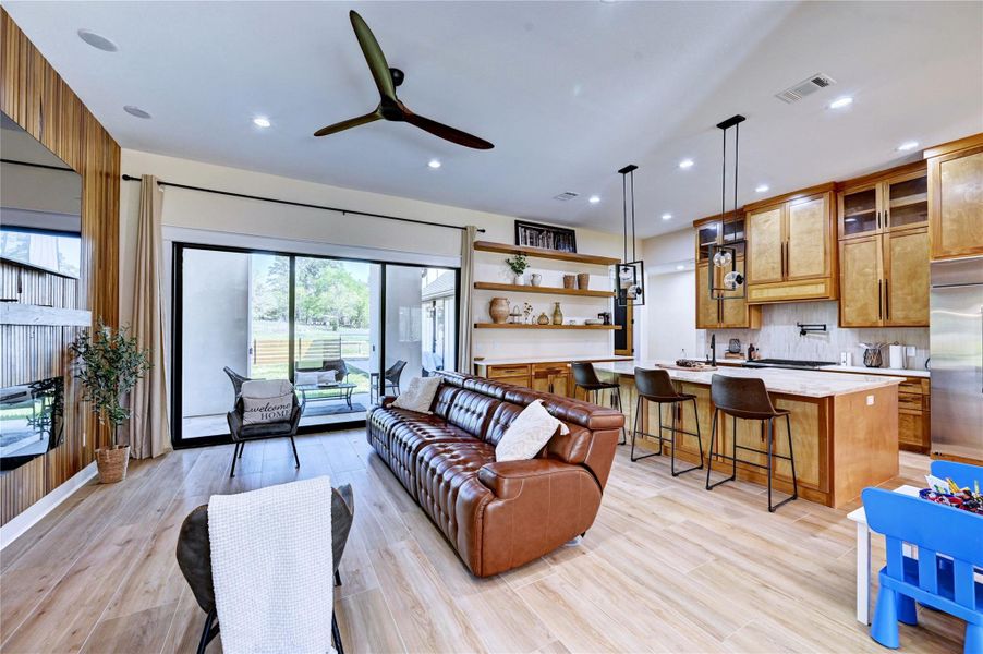 This spacious open-concept living area features a modern kitchen with wooden cabinets, an island with bar seating, and stylish pendant lighting. The cozy living space includes a leather sofa and large sliding glass doors that lead to an inviting outdoor patio, offering ample natural light and a seamless indoor-outdoor flow.
