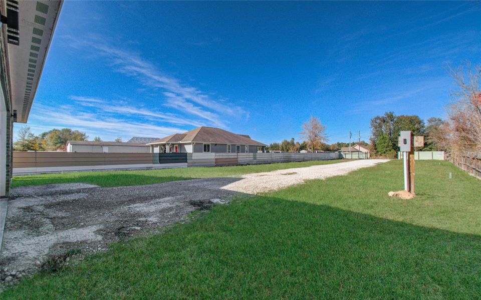 This photo shows a spacious yard with a gravel driveway and lush green grass. The property is fenced and features a modern house in the background, surrounded by trees, under a clear blue sky. Ideal for privacy and outdoor activities.