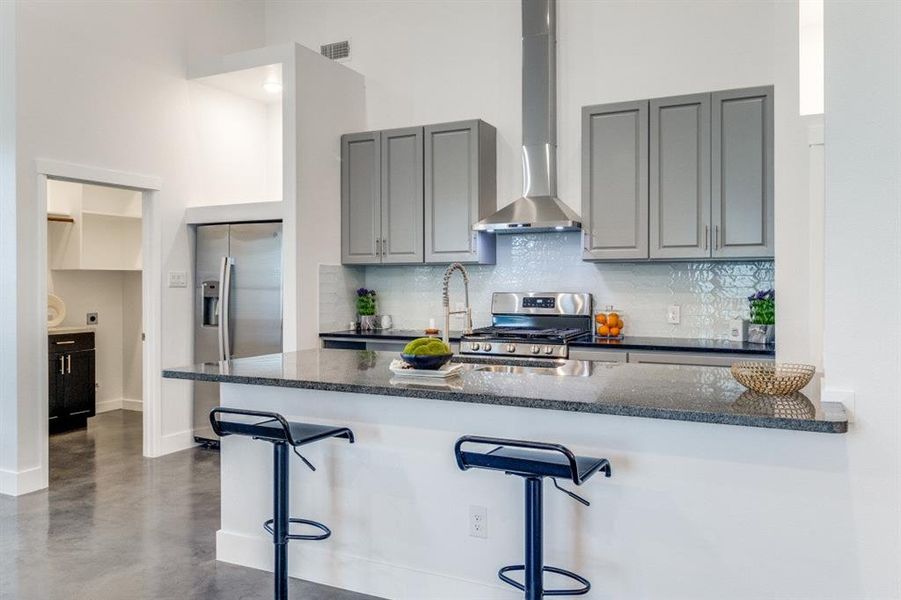 Kitchen featuring wall chimney range hood, kitchen peninsula, stainless steel appliances, a high ceiling, and a breakfast bar