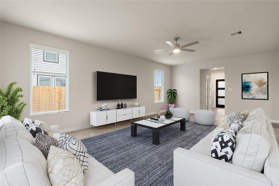This spacious living room features modern decor with a large sectional sofa, stylish cushions, and a sleek coffee table on a textured rug. Natural light fills the room through two windows, and the space is complemented by a wall-mounted TV, minimalistic stand, ceiling fan, and tasteful artwork.