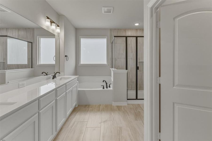 Spacious bathroom featuring a dual vanity with white cabinetry and light-toned countertops, a built-in tub, a separate shower with dark-framed glass enclosure, and light wood-look tile flooring Spacious bathroom featuring a dual vanity with white cabinetry and light-toned countertops, a built-in tub, a separate shower with dark-framed glass enclosure, and light wood-look tile flooring