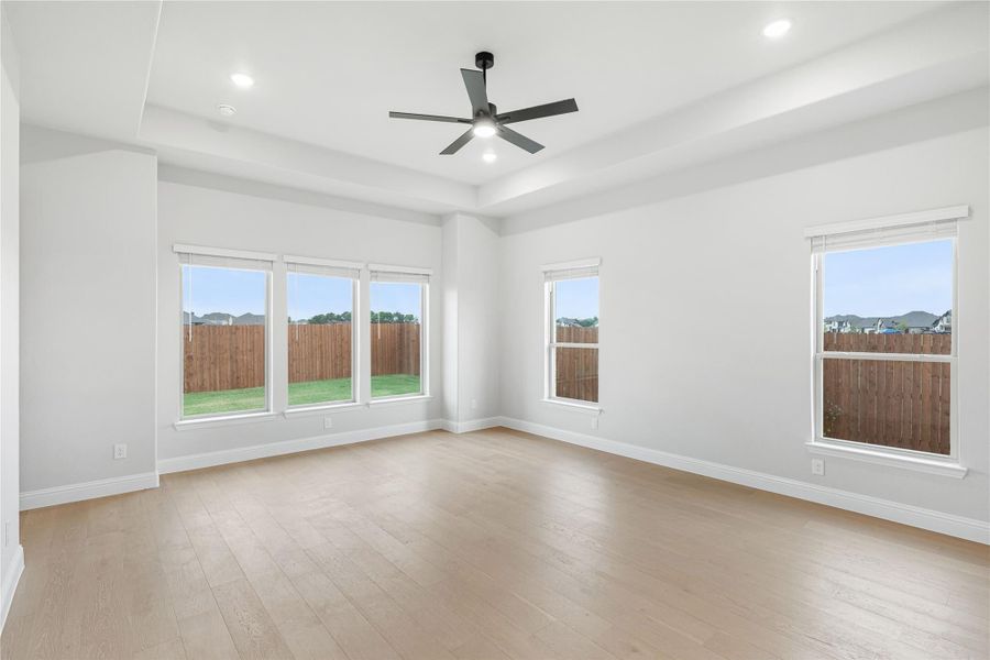 Empty room with tray ceiling, ceiling fan, light hardwood floors, and multiple windows overlooking backyard