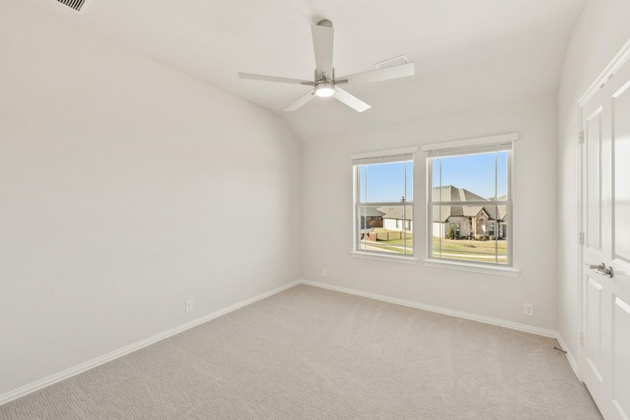 Empty bedroom with vaulted ceiling, ceiling fan, carpet flooring, and two windows overlooking neighboring homes