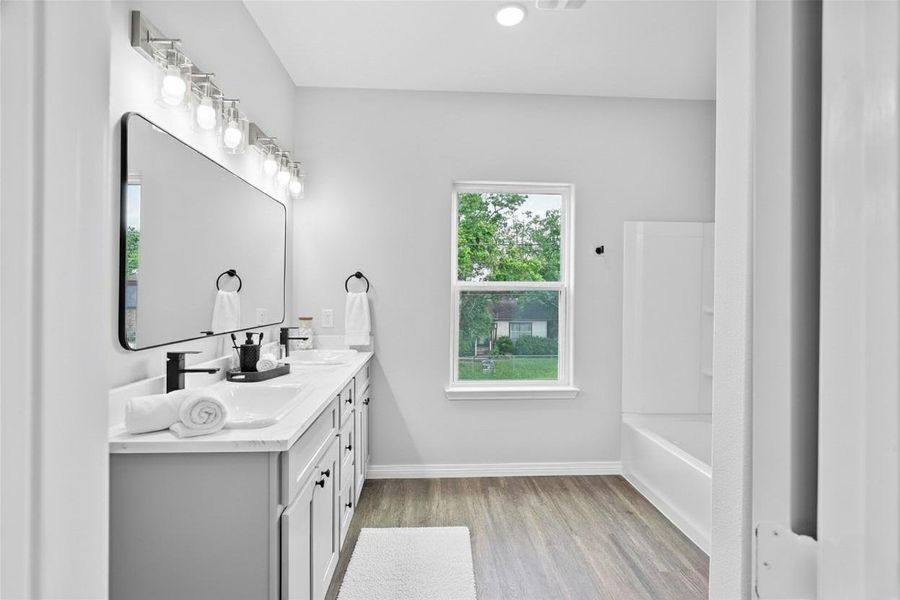 Bright, modern bathroom with dual sinks, large mirror, and ample lighting. Features a window with a view, sleek fixtures, and a clean design with wood-looking flooring. **Photo has been staged.**