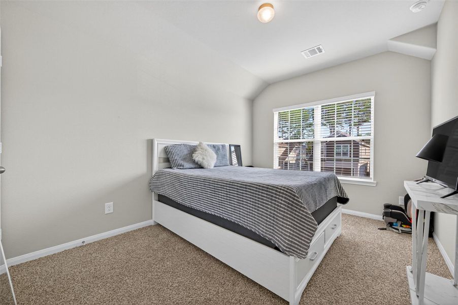 This bedroom features a cozy atmosphere with neutral walls and carpeted flooring. It includes a single bed with bedding, a window providing natural light, and a small desk with a monitor, making it ideal for relaxation or work.