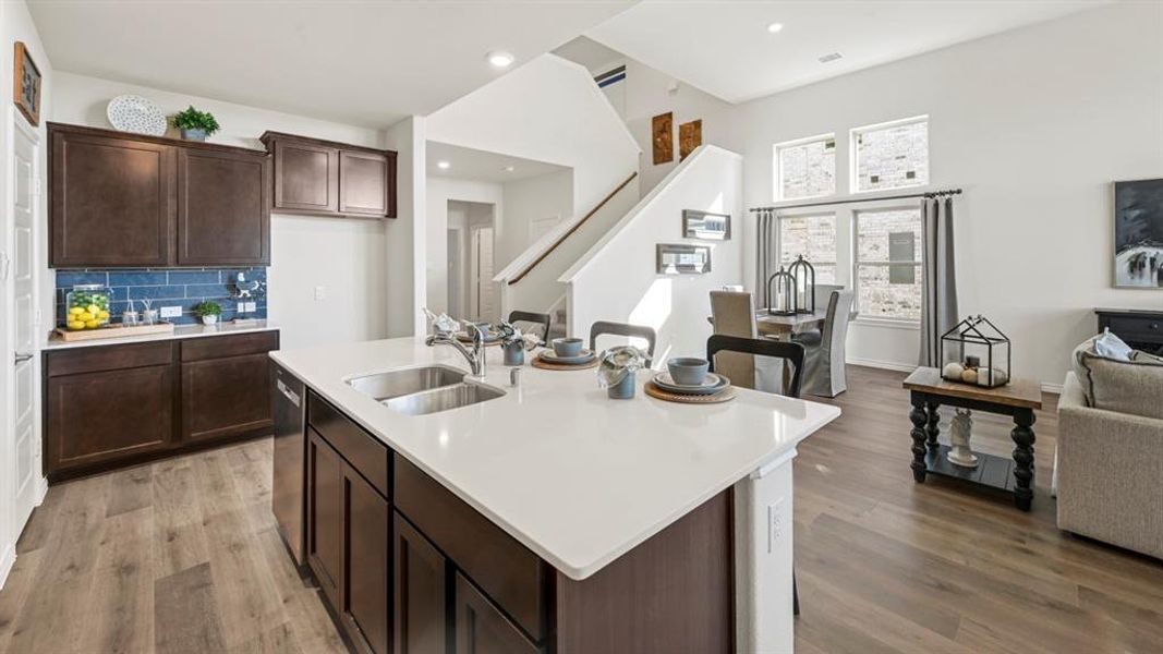 Open-concept living space featuring a central kitchen island with a double basin sink, rich wood-finish cabinetry, a blue subway tile backsplash, and wood-finish flooring