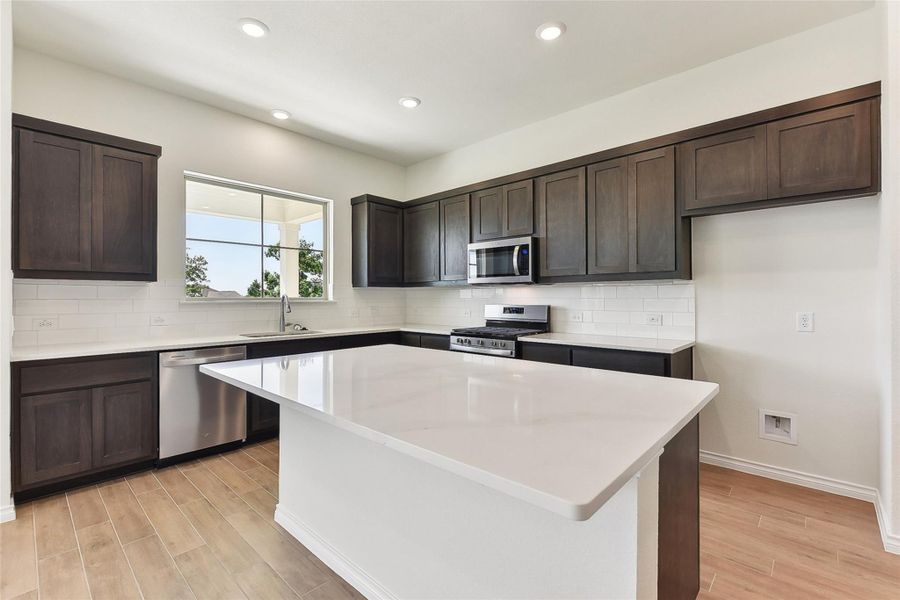 Kitchen featuring dark brown cabinetry, stainless steel appliances, backsplash, light wood-style flooring, and a kitchen island Kitchen featuring dark brown cabinetry, stainless steel appliances, backsplash, light wood-style flooring, and a kitchen island