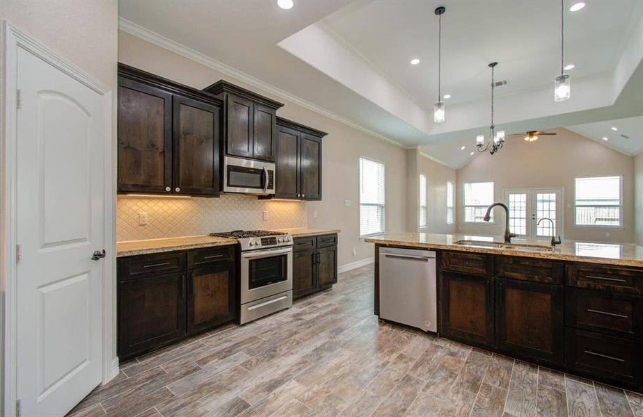 This is a spacious kitchen with dark wood cabinetry, stainless steel appliances, a central island with a sink, and modern lighting fixtures. The room features tiled flooring and is well-lit with natural light from several windows.