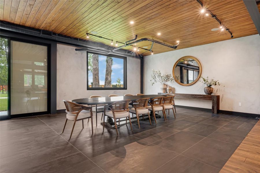 This elegant dining area features a spacious table with modern chairs, set against a backdrop of large windows offering scenic views. The room is enhanced by a wooden ceiling with sleek track lighting, a stylish mirror, and tasteful decor, creating a warm and inviting atmosphere.