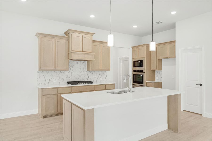 Kitchen with light wood cabinets, large center island with sink, marble backsplash, and stainless steel appliances.