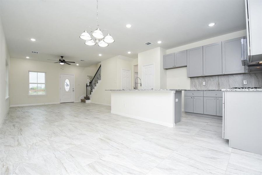 A spacious, modern kitchen with gray cabinets, marble countertops, and tile flooring, leading to a living area with a staircase and a front door.