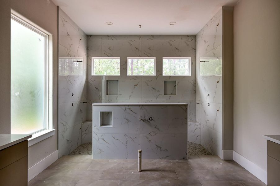 This photo showcases a spacious primary bathroom featuring elegant marble tile walls and floor. The room is brightened by a large frosted window and three smaller windows, providing natural light. There are built-in niches for ample storage.
