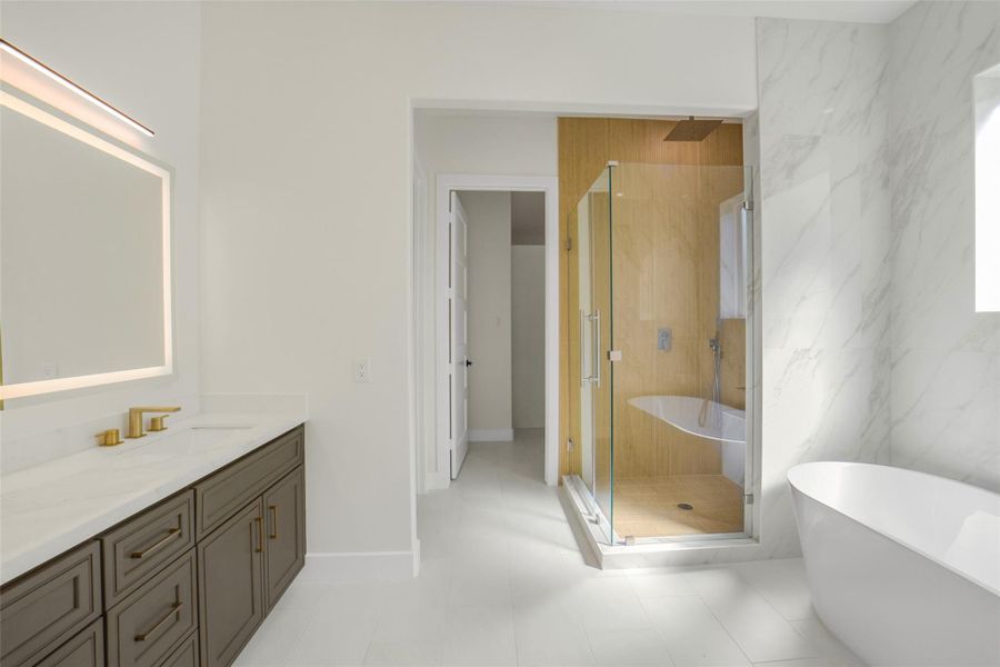 This modern bathroom features a spacious layout with a glass-enclosed shower, freestanding bathtub, and a wide vanity with elegant gold fixtures. The light color palette and large mirror enhance the airy feel.