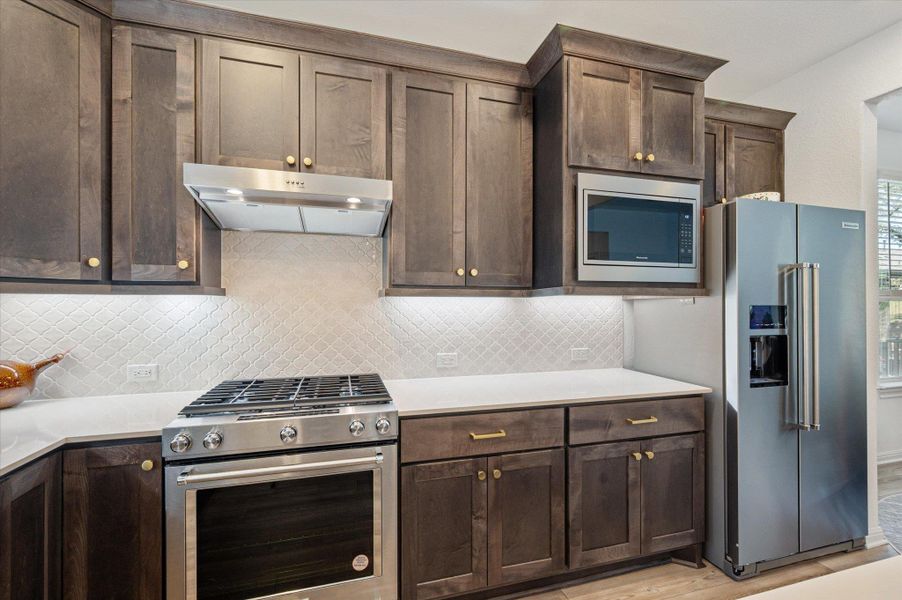Kitchen featuring stainless steel appliances, dark wood finish cabinetry, light wood finished floors, light stone countertops, and backsplash Kitchen featuring stainless steel appliances, dark wood finish cabinetry, light wood finished floors, light stone countertops, and backsplash
