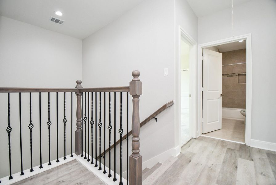 This photo shows a bright, modern stairway landing with wooden flooring and stylish iron railings. It leads to a bathroom with neutral tiles and a white door, creating a clean and inviting space.