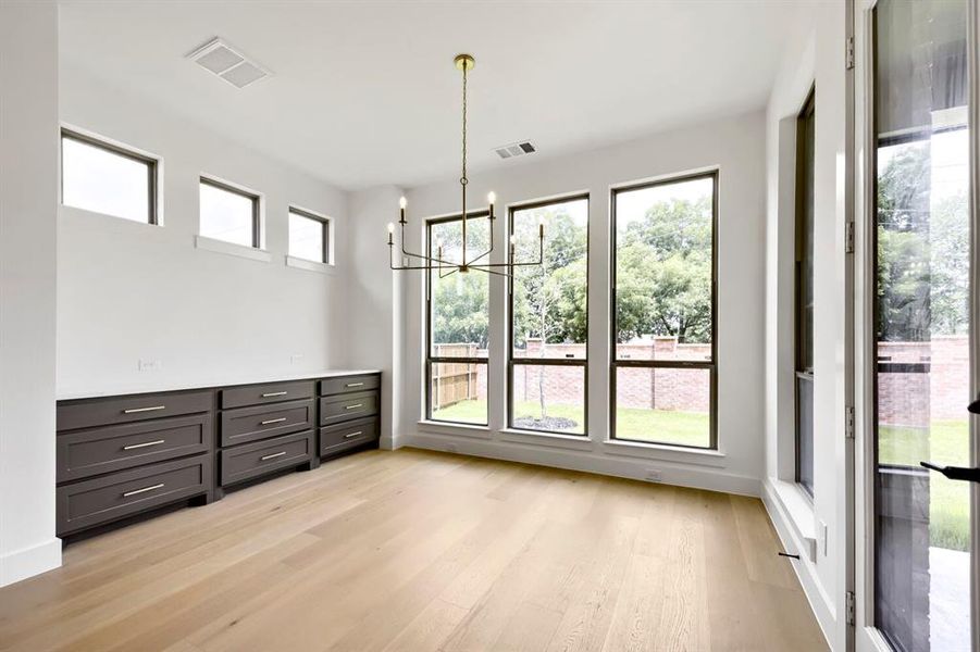 Spacious room featuring light wood-finish flooring, a contemporary chandelier, multiple large windows, and built-in dark gray cabinetry with a light countertop Spacious room featuring light wood-finish flooring, a contemporary chandelier, multiple large windows, and built-in dark gray cabinetry with a light countertop