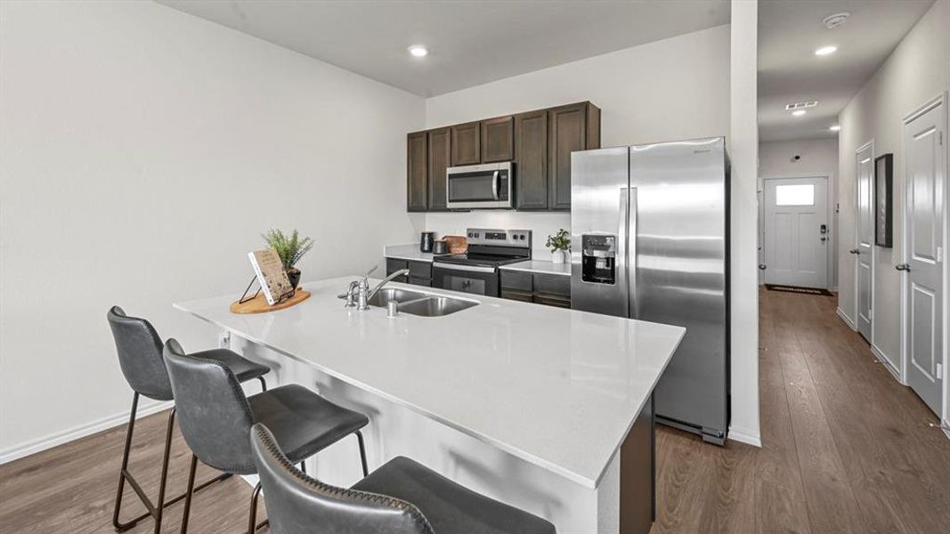 Kitchen featuring stainless steel appliances, dark brown cabinetry, dark wood-style floors, a breakfast bar area, and a center island with sink