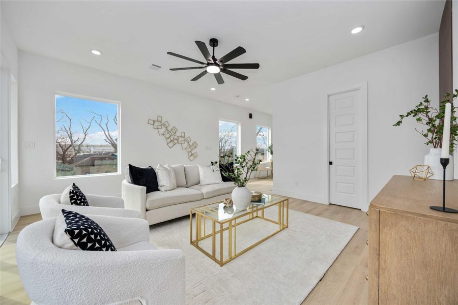 A bright, modern living room features neutral-toned furniture, a gold-accented glass coffee table, and large windows that fill the space with natural light.