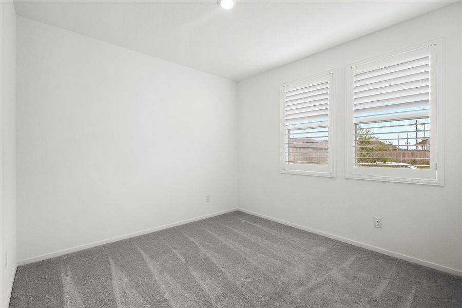 This bright room features clean white walls and soft gray carpeting. Two windows with shutters provide natural light and a view outside, creating an inviting and versatile space.