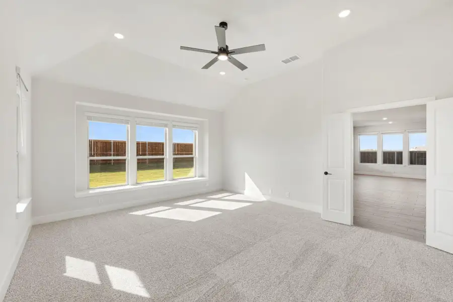 Empty bedroom with vaulted ceiling, ceiling fan, carpet flooring, and large windows overlooking a backyard